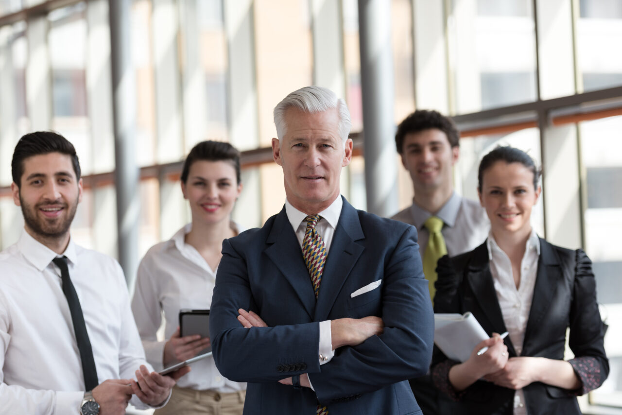 portrait of senior businessman as leader  at modern bright office interior, young  people group in background as team